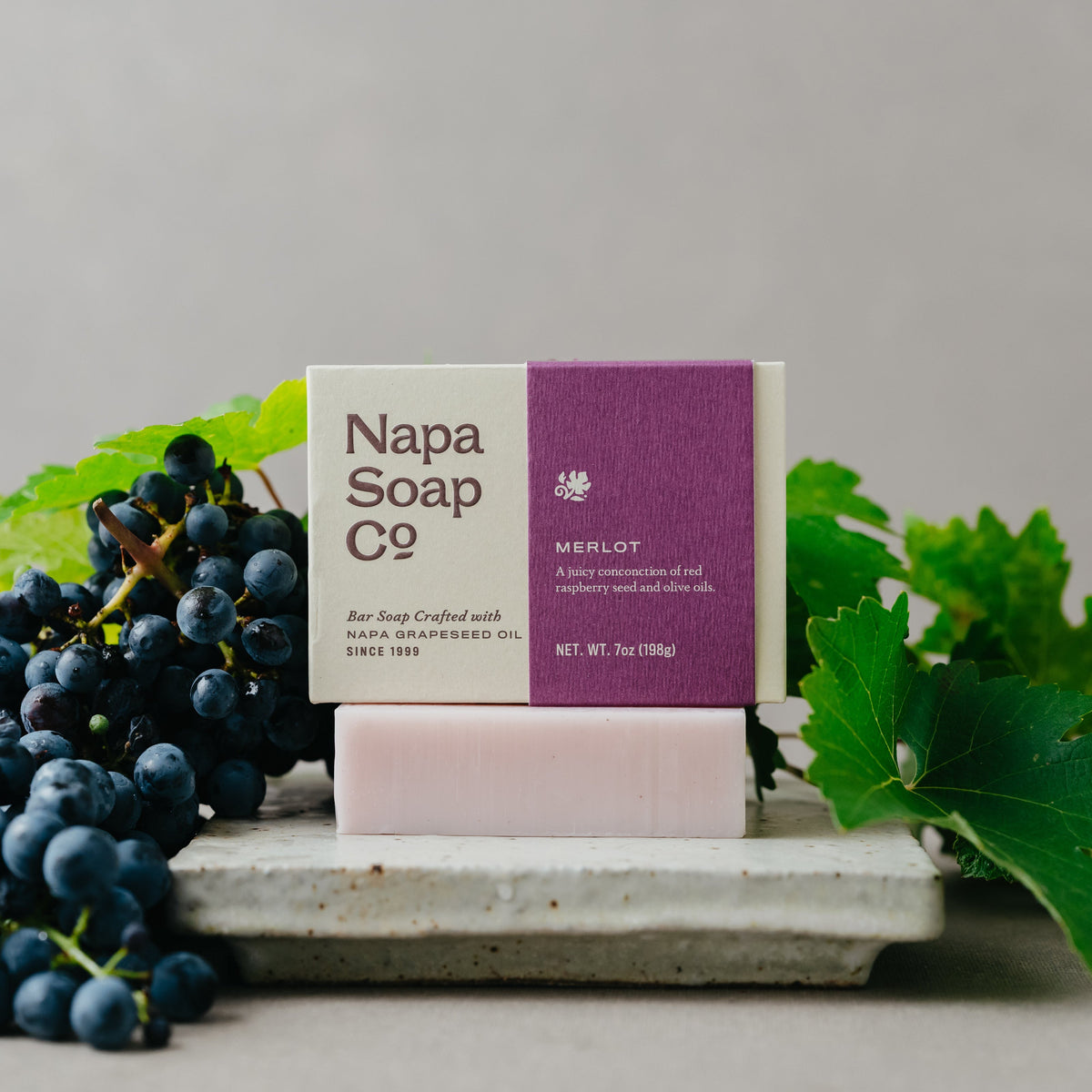 Napa Soap Company Merlot soap bar and packaging displayed with dark grapes and green grape leaves.